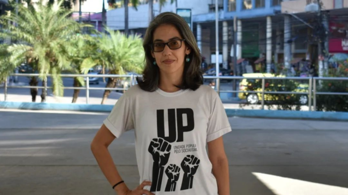 Na imagem, uma mulher branca, de óculos, cabelos brancos, usando uma camisa branca escrito UP na cor preta com vários punhos serrados desenhados. 