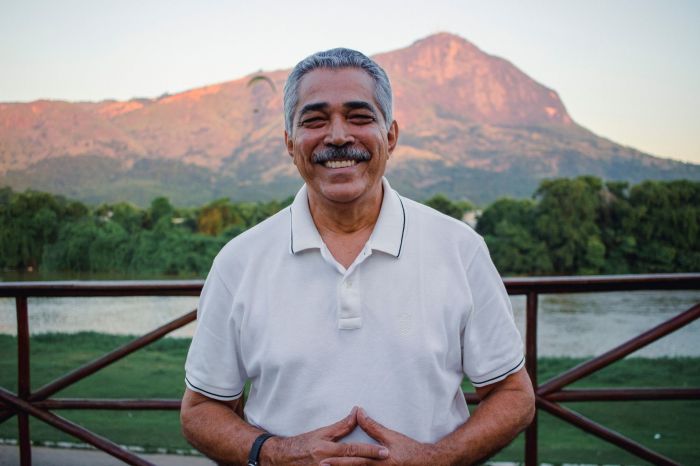 Léo Monteiro é um homem negro, de cabelo branco e bigode branco. Ele está em uma paisagem com morros altos, árvores e um rio ao fundo. Ele usa camisa polo branca e cruza as mãos.