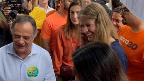 Mauro Tramonte, no canto esquerdo. Ele é um homem branco, de cabelo grisalho. Na imagem, usa uma camisa social azul clara com um adesivo verde, amarelo e azul. Ao lado, Luísa Barreto. Ela é uma mulher branca, loira e usa uma camisa social azul escura. Ao fundo, várias pessoas usando camisas laranjas, e uma usando uma camisa amarela segura uma câmera.