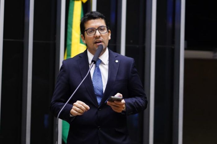 Marcelo Queiroz é um homem branco de cabelo preto e óculos de grau. Na imagem, ele usa terno azul escuro, gravata azul e camisa branca. 