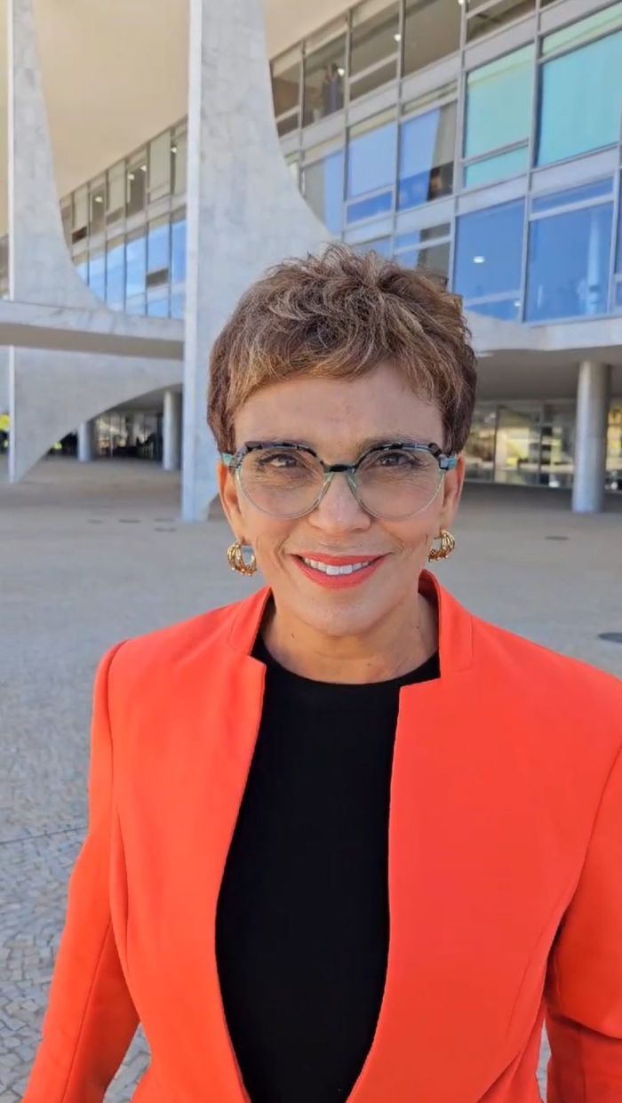 Marília Campos é uma mulher branca, de cabelos claros e curtos. Na imagem, ela usa camisa preta, blazer laranja, batom laranja, brincos dourados e óculos de grau.