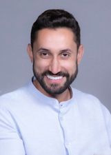 Paulo Bigodinho é um homem branco, de barba e cabelos castanho escuro, usa óculos de grau com alça preta, e na imagem ele sorri enquanto usa uma camisa social branca clara.