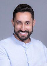 Paulo Bigodinho é um homem branco, de barba e cabelos castanho escuro, usa óculos de grau com alça preta, e na imagem ele sorri enquanto usa uma camisa social branca clara.