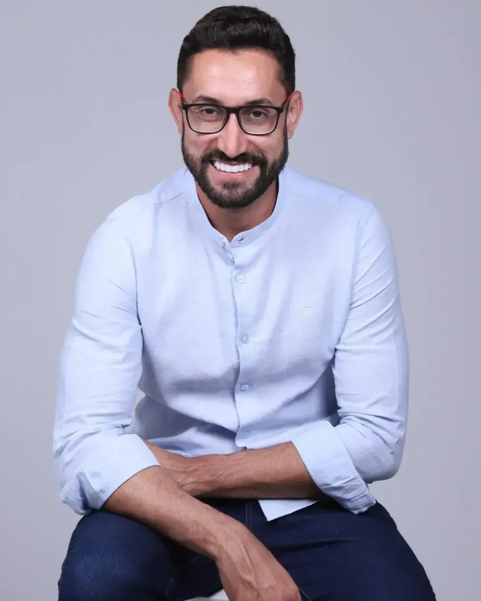 Paulo Bigodinho é um homem branco, de barba e cabelos castanho escuro, usa óculos de grau com alça preta, e na imagem ele sorri enquanto usa uma camisa social branca clara.