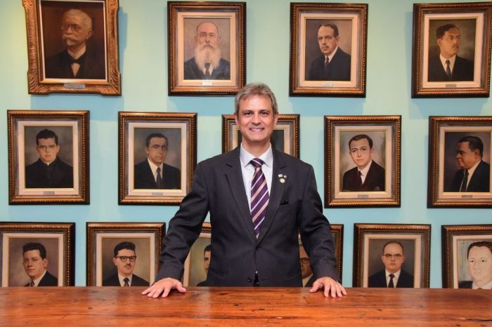 Homem branco, de cabelo branco, terno preto e gravata listrada parado com as mãos em uma mesa de madeira. 