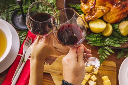 Pessoas brindam com taças com vinho tinto. Ao fundo, uma mesa com petiscos