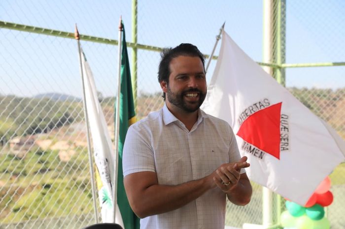 Túlio Raposo é um homem branco, de cabelos castanhos escuros e barba da mesma cor. Na imagem, a bandeira de Minas Gerais balança no fundo, ao lado da bandeira do Brasil. 