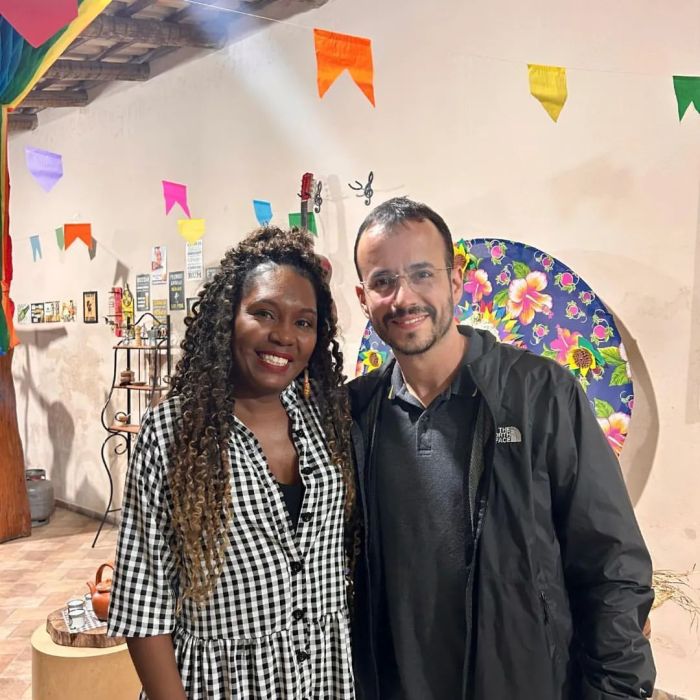Na imagem, uma mulher preta com cabelos castanhos com pontas loiras usa uma camisa xadrez preta e branca. Ao lado dela, um homem branco de óculos, cabelo preto, barba e camisa cinza e casaco preto. Ao fundo deles, tem bandeirinhas coloridas de festa junina e uma parede bege.