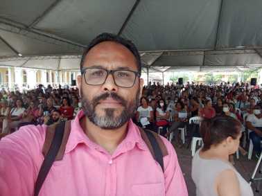 Wanderson Rocha é um homem preto, de cabelo cinza e barba cinza/branca. Na imagem, ao fundo tem várias pessoas sentadas em cadeiras de plástico. Wanderson usa um óculos de grau com aro preto, mochila com alça marrom e preta e uma camisa de botão rosa.