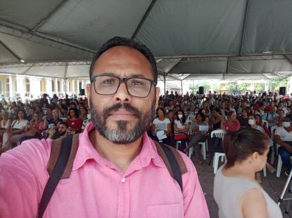 Wanderson Rocha é um homem preto, de cabelo cinza e barba cinza/branca. Na imagem, ao fundo tem várias pessoas sentadas em cadeiras de plástico. Wanderson usa um óculos de grau com aro preto, mochila com alça marrom e preta e uma camisa de botão rosa.