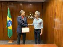 Dois homens brancos de pé, usando roupas sociais, apertam as mãos ao lado da bandeira do Brasil, com uma parede de madeira ao fundo.