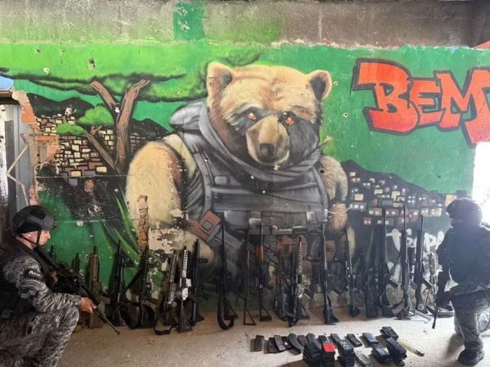 Uma parede exibe o grafite de um urso gigante vestindo colete à prova de balas e empunhando um fuzil 