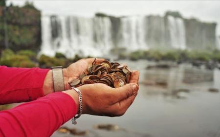 Homem segura moedas enferrujadas com as mãos. No fundo, as Cataratas do Iguaçu