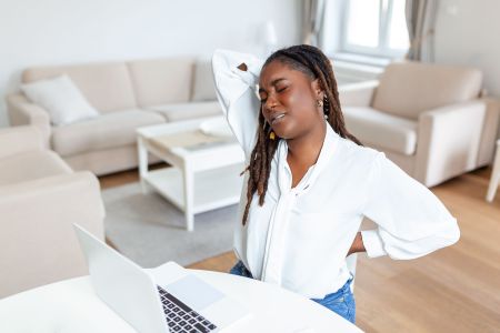 Young African businesswoman having back pain while sitting at office desk. Businesswoman Holding Her Back While Working On Laptop At Office Desk