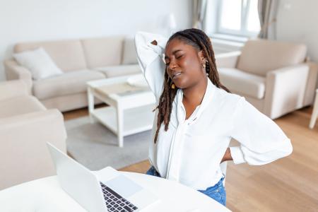 Young African businesswoman having back pain while sitting at office desk. Businesswoman Holding Her Back While Working On Laptop At Office Desk