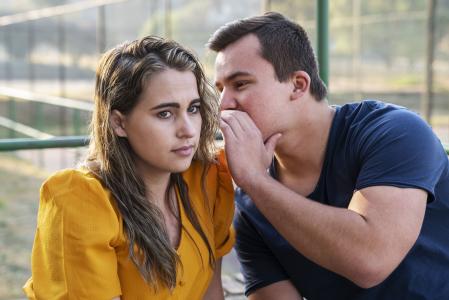 Homem branco com blusa azul contando um segredo para uma mulher branca e de cabelos castanhos e blusa amarela