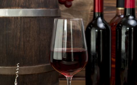 Taça com vinho tinto e garrafas da bebida no fundo, com também um barril