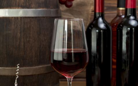 Taça com vinho tinto e garrafas da bebida no fundo, com também um barril