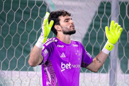 Goleiro do Cruzeiro com uniforme roxo e luvas verde-limão, em pé na frente da rede, com as mãos levantadas e olhos fechados em um gesto de oração ou agradecimento.