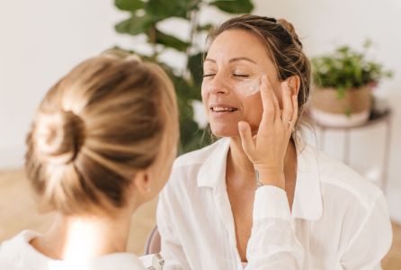 Garota loira e caucasiana aplicando creme anti-idade no rosto da mãe. A jovem está de costas para a câmera, enquanto a mãe aparece sorrindo e na direção da câmera