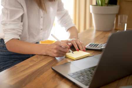 Mulher de camisa branca escevendo em bloco de anotações, com calculadora ao seu lado e notebook à sua frente enquanto utiliza sua conta digital.