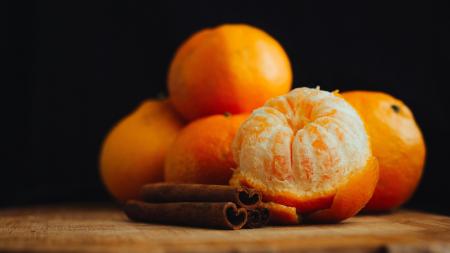 Cinco tangerinas empilhadas, sendo uma descascada, e paus de canela em cima de uma mesa de madeira e um fruto preto.