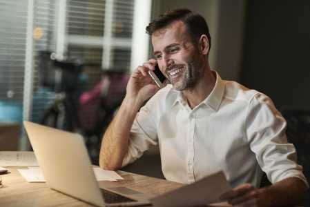Homeme de camisa branca sorrindo enquanto fala no telefone e olha para tela do notebook que está na frente dele, em cima de uma mesa.