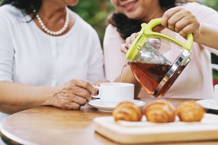 Close de mulheres sentadas, na frente delas uma mesa com café e um bule com chá. Elas sorriem