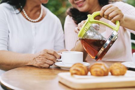 Close de mulheres sentadas, na frente delas uma mesa com café e um bule com chá. Elas sorriem