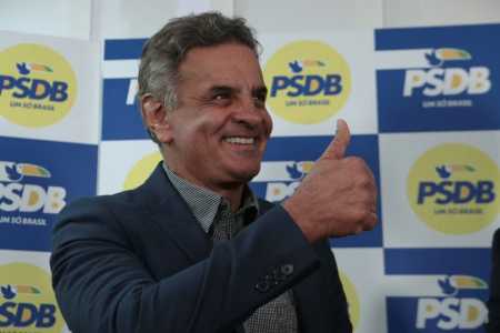 Aécio Neves em terno azul escuro sorrindo e fazendo joia com a mão com painel do PSDB de fundo em amarelo e azul