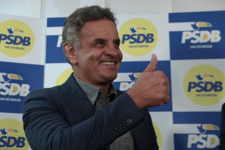 Aécio Neves em terno azul escuro sorrindo e fazendo joia com a mão com painel do PSDB de fundo em amarelo e azul