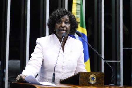 Benedita da Silva é uma mulher negra com cabelos cacheados, curtos e escuros. Veste camisa branca.