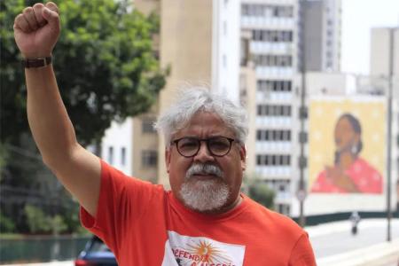 Edmilson Costa é um homem branco com cabelos e barba branca, utiliza óculos pretos e camisa vermelha com o braço levantado. Ao fundo, prédios e árvores.