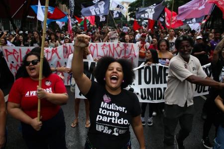 Samara está no meio da imagem e é uma mulher preta com blusa preta escrito Mulher Tome Partido com o braço para cima e com a boca aberta gritando