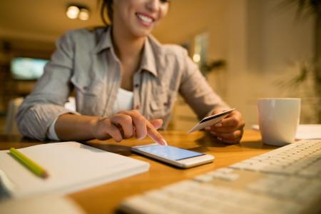 Mulher usa celular para serviços financeiros enquanto segura cartão de crédito, com braços apoiados em cima de uma mesa e um caderno ao lado.