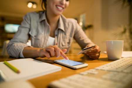 Mulher usa celular para serviços financeiros enquanto segura cartão de crédito, com braços apoiados em cima de uma mesa e um caderno ao lado.