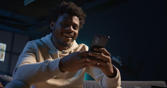 Homem com casaco de moletom sorrindo enquanto digita em celular com as duas mãos.