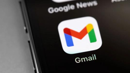 Close-up da tela de um smartphone exibindo o ícone do aplicativo Gmail sobre um fundo escuro. O logotipo colorido em formato de "M" aparece centralizado, com a palavra "Gmail" logo abaixo. Na parte superior da tela, é possível ver parcialmente o nome do aplicativo Google News, enquanto a lateral do aparelho também está visível.
