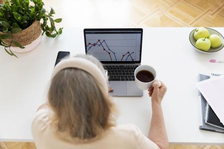Mulher acompanha gráficos de desempenho dos seus investimentos pelo notebook enquanto toma uma xícara de café.