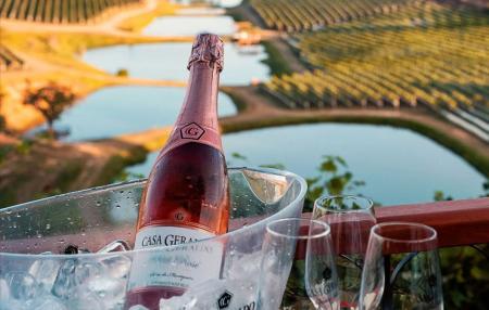 Garrafa de espumante rosé em um balde transparente com gelo, acompanhada por taças sobre uma varanda com vista para vinhedos. Ao fundo, lagos artificiais refletem a luz dourada do entardecer, criando uma paisagem rural associada ao enoturismo e à degustação de vinhos.