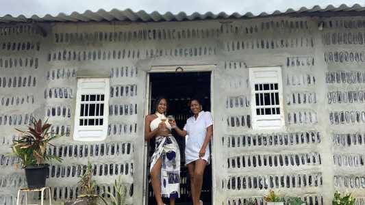 Duas mulheres sorriem enquanto brindam com taças na entrada de uma casa construída com garrafas de vidro incorporadas às paredes. A fachada cinza exibe fileiras de garrafas visíveis entre a argamassa, além de duas janelas brancas e um telhado de fibrocimento. Vasos com plantas decoram a frente da residência, destacando a proposta de construção sustentável com materiais reciclados.