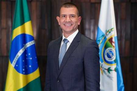 Douglas Ruas é um homem branco de cabelo raspado. Veste terno preto com camisa branca e gravata azul clara. No fundo, parede de madeira com bandeira do Brasil e do Rio.
