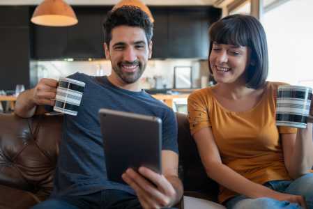 Um homem de camisa azul e uma mulher de blusa amarela, sentados lado a lado, sorriem enquanto usam um tablet para acessar serviços financeiros.