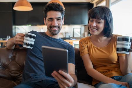 Um homem de camisa azul e uma mulher de blusa amarela, sentados lado a lado, sorriem enquanto usam um tablet para acessar serviços financeiros.