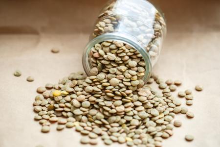Close-up de um pote de vidro tombado sobre uma superfície de madeira, com lentilhas secas espalhadas ao redor. Os grãos, em tons de verde, bege e marrom-claro, saem da abertura do recipiente, destacando sua textura e aparência natural em um ambiente bem iluminado.