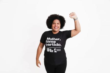 Samara Martins é uma mulher negra em fundo branco com camisa preta escrita Mulher Tome Partido e calça preta. Seu braço está levantado para cima.