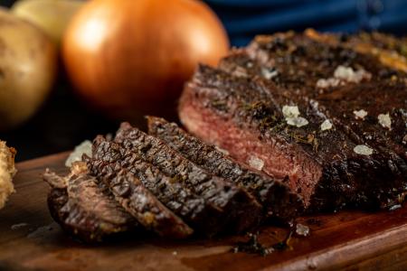 Close-up de uma peça de carne bovina grelhada sobre uma tábua de madeira, parcialmente fatiada e com interior rosado. A carne está temperada com sal grosso e ervas, enquanto cebolas e batatas desfocadas aparecem ao fundo, compondo uma cena que remete a um churrasco.
