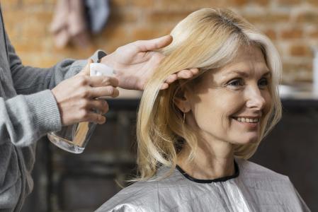 Mulher loira de meia-idade sorri enquanto recebe cuidados no cabelo. Uma pessoa ao lado segura uma mecha de seus cabelos e borrifa um produto com um frasco spray transparente. A mulher usa uma capa de salão prateada, e o ambiente tem uma parede de tijolos desfocada ao fundo, sugerindo um salão de beleza ou espaço de cuidados pessoais.