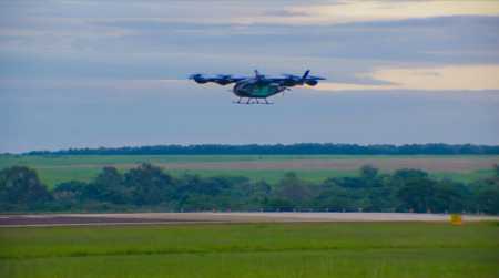 Drone de grande porte em voo sobre uma pista de pouso e decolagem, com áreas verdes e árvores ao fundo sob um céu nublado. A aeronave está em destaque no centro da imagem, próxima da pista.