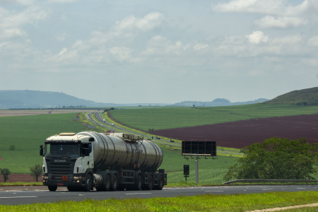 Caminhão-tanque trafega por rodovia cercada por lavouras em área agrícola, com campos cultivados e colinas ao fundo.